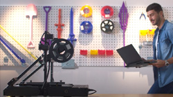 A person holding a laptop is approaching a 3d printer on a workbench from the right. A white pegboard is in the background.