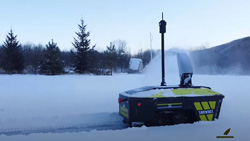 A black and yellow wheelbarrow-sized vehicle has a mast twice its height protruding from the top and a chute in the front that is blowing snow away from the camera. A clear path through the snow can be seen behind the vehicle.