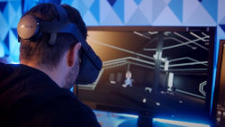 a man with a VR headset on with a hockey puck sized device in the back that holds the neuro sensors. He is looking at a monitor that is displaying a VR environment