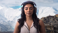 A person with long brown hair is wearing a headphone-like device on their head facing the camera. Their eyes are closed. There is a snowy mountain scene behind them.