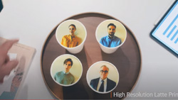 A top view of four paper coffee cups on a dark brown round platter that is sitting on a table. Four different images of people appear at high-resolution in the foam of the lattes.