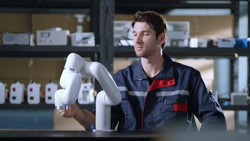 A person in a blue jacket has their hand on a desktop, white, robot arm with 9 joints.