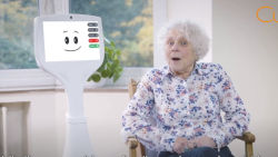 an elderly woman sitting next to a tall, thin, rolling, white, companion care robot with a smiling clipart face