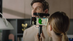 A view from over the left shoulder of a person holding a selfie stick with a phone and a pill-shaped 3d scanner horizontally attached. On the phone's screen is a 3d representation of the person they are scanning. The person being scanned is looking away from the camera and appears like they are trying to stay motionless.