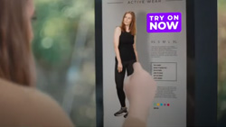 A view from behind and to the right of a person with one finger pointed near a vertical screen. The screen has an image of a clothing ad that has text Try On Now.