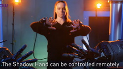 A front view of a person holding their hands open in front of them. Glove-like attachments are on both hands. Two robot hands can be seen in the foreground mimicking the human's hands. The test reads The Shadow Hand can be controlled remotely.