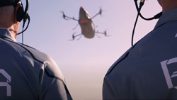 A view between the shoulders of two people with headsets on, of an eVTOL in the air. The eVTOL is shaped like an elongated tear drop with 2 propellers on struts in front and back.