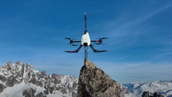 A black and white flying robot drone sits on top of a high mountain peak.