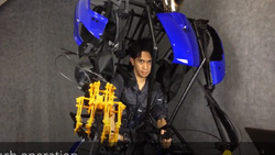 A view of a person in an open framed large exoskeleton. This exoskeleton extends away from the body to make it appear larger. The exoskeleton hand is yellow and the chest panels are blue.