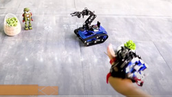 A POV camera angle of a person's hand with a device that is attached like a glove and is pointing at a small blue track robot on the floor. The robot has an arm with pinchers attached to the top of the tank-like chassis that makes it about a foot in height.