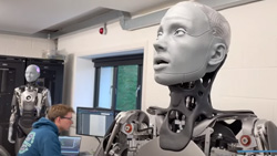 In the right foreground a light gray life-like humanoid robot is seen from the shoulders up. It is 'looking' to the right with the mouth slightly open as if in awe. In the background is a person in a blue hoodie at a computer in front of a small window. Further back is another standing humanoid robot identical to the one in the foreground