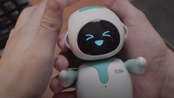 A close-up of a palm-size white and cyan robot in a person's hands. It has an LCD screen for a face with cyan eyes and mouth showing a happy face