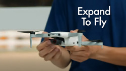 A close-up of a person holding a white drone with both hands (looks about 12 inches across). The text reads Expand to Fly.