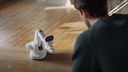 A view from behind left of a person kneeling in front of a white and black robot arm on that is on a wood floor.