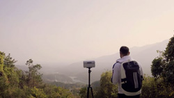 A person with a black backpack and white jacket is facing away from the camera looking at a foggy valley. There is a small white box on top of a tripod next to him.