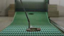 A closeup of a putter hitting a ball on a practice putting green that slopes up at the end where the hole is.