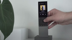 A person is placing a gray and black remote control with a small screen at the top in a docking station.