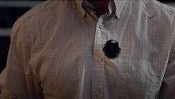 A closeup shows the front of person in a white button-down shirt with a small black oval device near the left pocket.