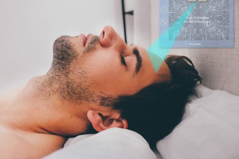 A person is laying on a bed with their eyesclosed. A blue beam is being directed at their forehead from a screen on the headboard.