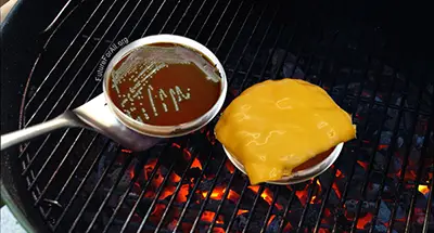 Two petris dishes on a grill with hot coals, one dish has cheese melted on it, the other is being flipped with a spatula