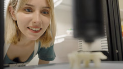 A closeup of a person, indirectly facing the camera, leaning in and looking at a 3d model being printed. The person is in focus, the rest of the image is blurred.