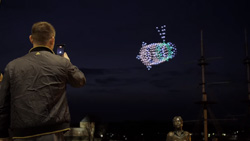 A person with their back to the camera is holding up a smartphone at night towards a city skyline with lighted drones in the air in the shape of a satellite