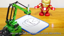 A tabletop green robotic arm holding a black marker sits in front a dry erase board that has a line drawing of the face of a toy Iron Man figure that sits in front of it.