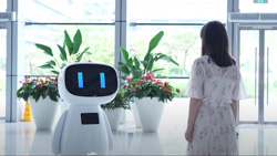In a brightly lit glass building a person stands facing a white robot with two blue vertical lines as eyes. The robot is chest high to the person. The robot's head is shaped like a rounded rectangle on its side with a big black screen.