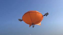 An orange balloon shaped like a MandM candy with fins on the back and a propellor on each side and a camera mounted underneath in the blue sky is viewed at an angle from below