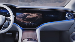 A view from the back seat of a vehicle dashboard that is one big display and perhaps a touchscreen with an air vent on the right side. A picture of rolling hills is in the center. There is blue LED trim around the bottom of the dashboard. The interior of the sedan is white including the steering wheel.
