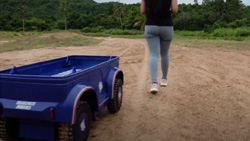 A person is walking away from the camera on a dirt road. A blue 4-wheeled cargo robot follows behind