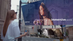 A view from behind and to the right of a person on a city street during the day with AR glasses on viewing a large semi-transparent screen showing a movie