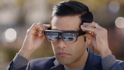 a closeup of a person putting on black-rimmed smart glasses with thick lenses. A wire is hanging from the back left side of the glasses.