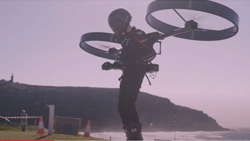 A hazy image of a person in an all black flight suit with a motorcycle helmet on. The have two drone-like propellers facing downward strapped to their back. The location is the sandy shoreline of a body of water.