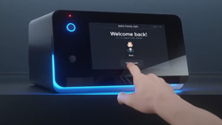 A closeup of a person with their index finger extended towards a black box with a touchscreen on the front. The box is about the size of a loaf of bread. It has a round blue button in the top left and has blue LED lights bordering the bottom. The screen says Welcome Back!