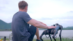 A rear view of a person sitting with a small four-legged robot at the edge of a lake.