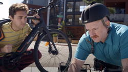 A view of two people outside during the day. The person on the left, with a yellow striped shirt, is kneeling with their head and shoulders through the middle of a mountain bike as if they are carrying it, and is facing the other person. The person on the right, in a light blue polo shirt and bike helmet, looks annoyed and is staring at the ground.