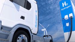 A closeup of a semi-truck pulled up to a hydrogen fuel pump. A wind turbine is in the background.