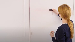 A view from behind shows a person holding a small black device against a wall with their right hand and holding a pencil with the other. A red laser line on the wall is being emitted from the device and is leading away from the user