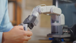 A closeup of a small white and gray robot arm grabbing something the size of a thumb drive from a person's right hand.