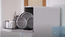 A white, compact dishwasher on a kitchen countertop is open and has dishes loaded. It is about the size and shape of a large tower PC.