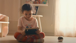 A closeup of a child about 4 years old sitting cross-legged on the floor holding and looking at a tablet with a small ball-shaped black and white robot