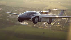 A white and black sports car with wings and a spoiler in back is in the air high above the countryside