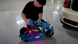 A person in a parking garage is kneeling behind a beige sedan with the trunk open. The person is folding a blue scooter into a smaller form.