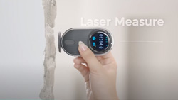 A person is holding a gray, slim, pill-shaped device with an LCD display to a drywall panel.
