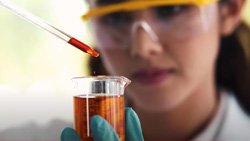 A closeup of a person with safety goggles on and wearing blue rubber gloves using a pipette to put a red liquid into a 3/4 full small beaker.