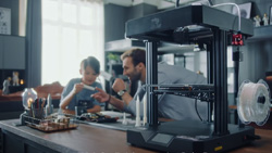 A 2 foot tall tabletop black 3d printer is in the foreground sitting on a wood worktable. The printer is open-framed with square platforms on top and bottom and 3 supporting posts in between, 2 in front and one in back. The printing platform is raised half-way. An adult and child are in the background looking at a 3d print.