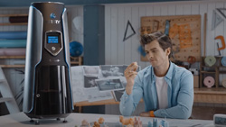 A person is sitting at a table looking at a small tan object in their hand. A tall, silver and black, cylinder-shaped 3d printer is on the table to the left. A small LCD display can be seen on the front of the printer.