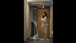 a person in a robe and holding a cup is standing outside their room facing a waist high, cylindrical-pedestal-shaped, silver and black robot. The person is looking at the top of the pedestal which presumably is the user interface