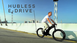 A person is riding a carbon black bicycle over a bridge on a sunny day. The bike has no hub or spokes on either wheel.